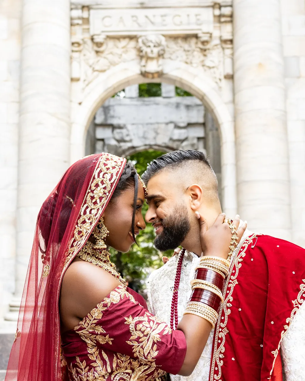 Bride in red with chooda touching foreheads with the groom under a stone arch