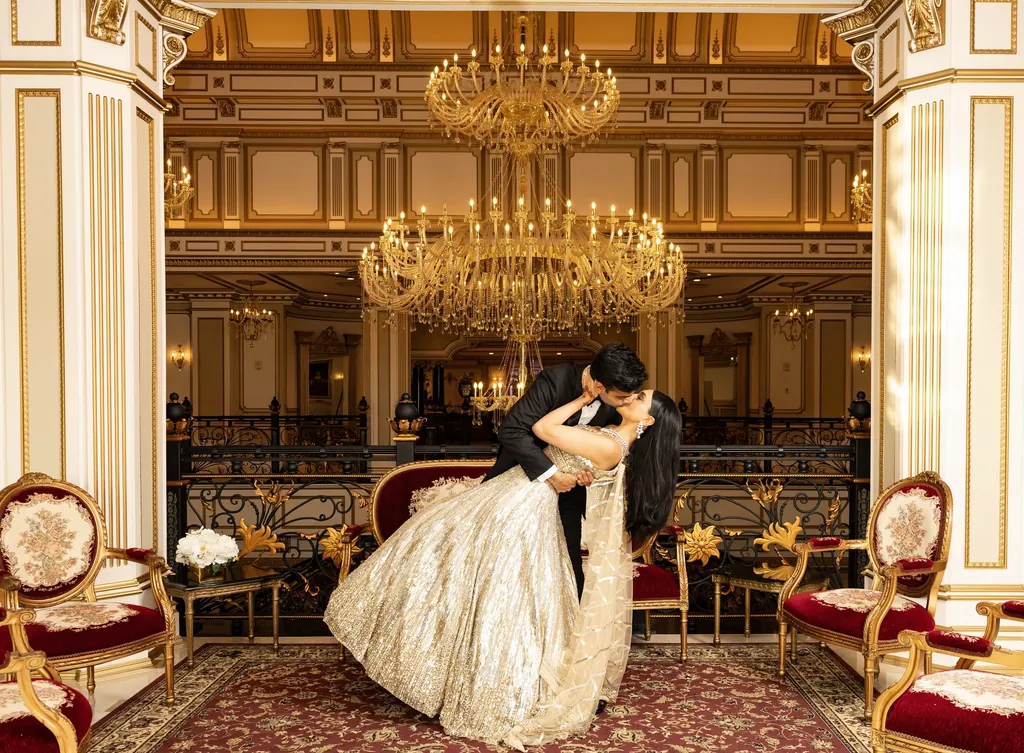 Groom dips the bride for a kiss in a gilded ballroom under chandeliers