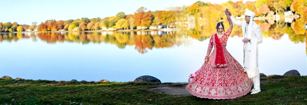 Bride in a red lehenga twirls as the groom in an ivory sherwani lifts her hand, beside an autumn lake