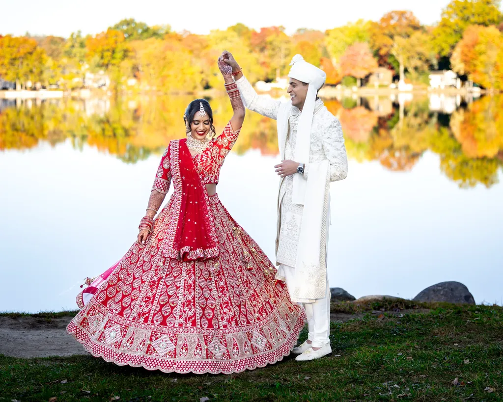 Bride in a red lehenga twirls with the groom beside an autumn lake