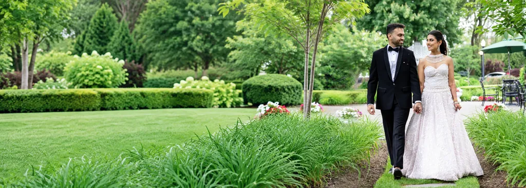 Groom in a black tuxedo and bride in a beaded white gown and Indian jewellery walking a garden path