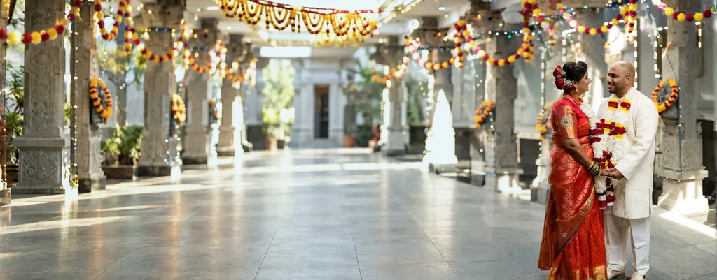 Bride in a red silk saree and groom in ivory with flower garlands, in a temple corridor strung with marigolds