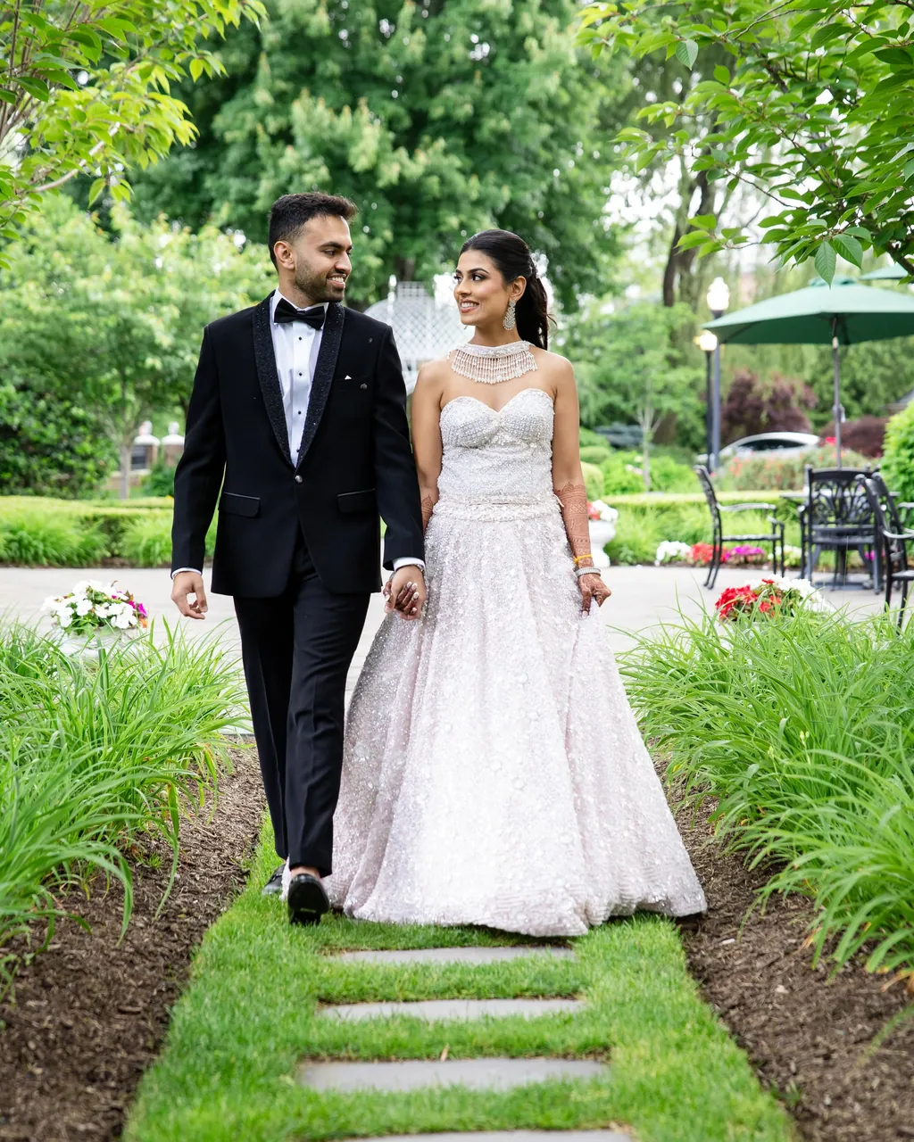 Bride in a beaded gown and groom in a tuxedo walking a garden path