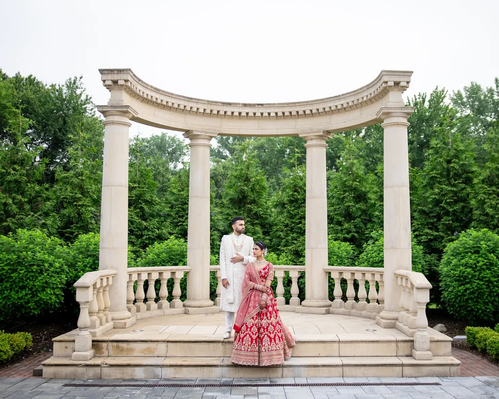 Bride in red and groom in ivory at a columned garden pavilion