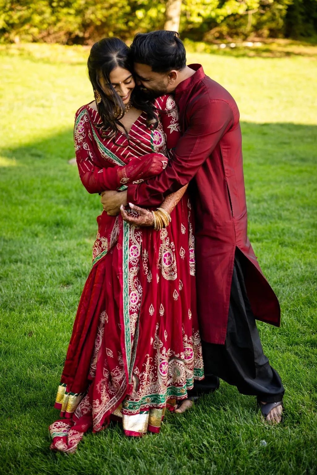 Groom in burgundy embracing the bride in a red lehenga on a lawn