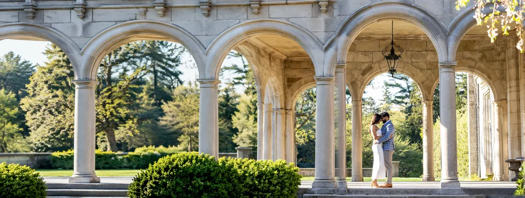 Couple embracing under a sunlit stone arcade with a hanging lantern, estate gardens beyond