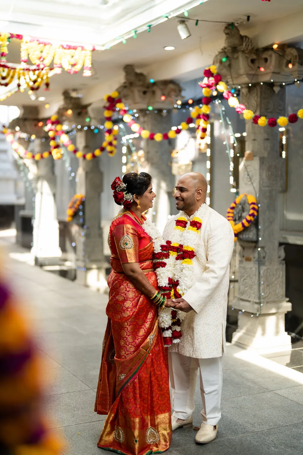 Bride in a red silk saree and groom with flower garlands in a temple corridor