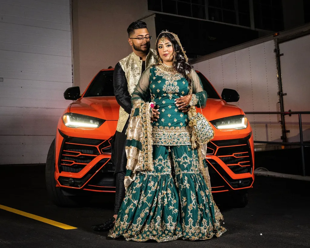 Couple in emerald wedding outfits beside an orange sports car at night