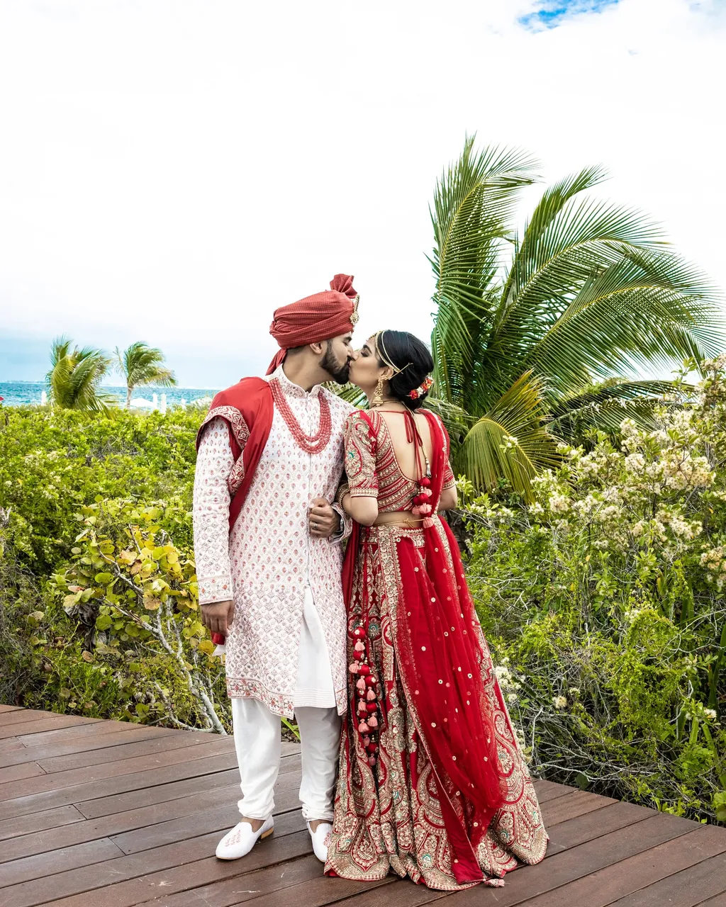 Bride in a red lehenga and groom in a red turban on a seaside boardwalk