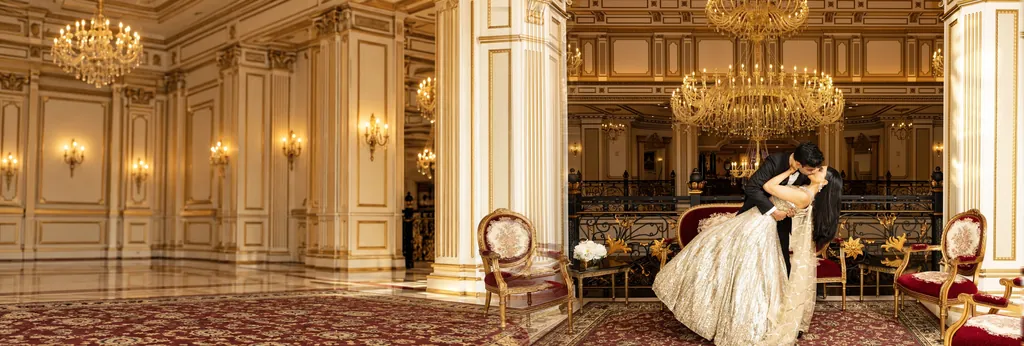 Groom dips the bride for a kiss in a gilded ballroom under crystal chandeliers
