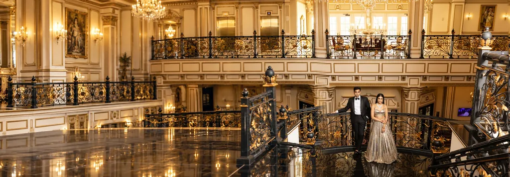 Bride in a silver lehenga and groom in a tuxedo on a gilded wrought-iron staircase in a grand hall