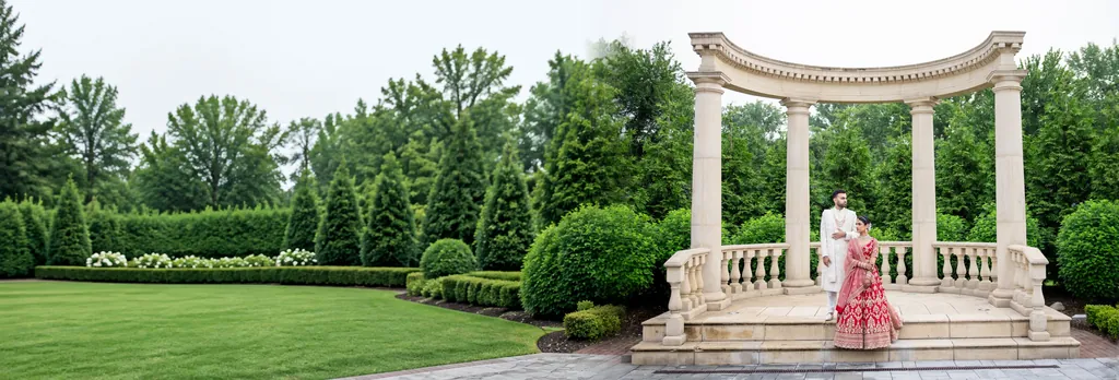 Bride in a red lehenga and groom in ivory under a columned garden pavilion on an estate lawn